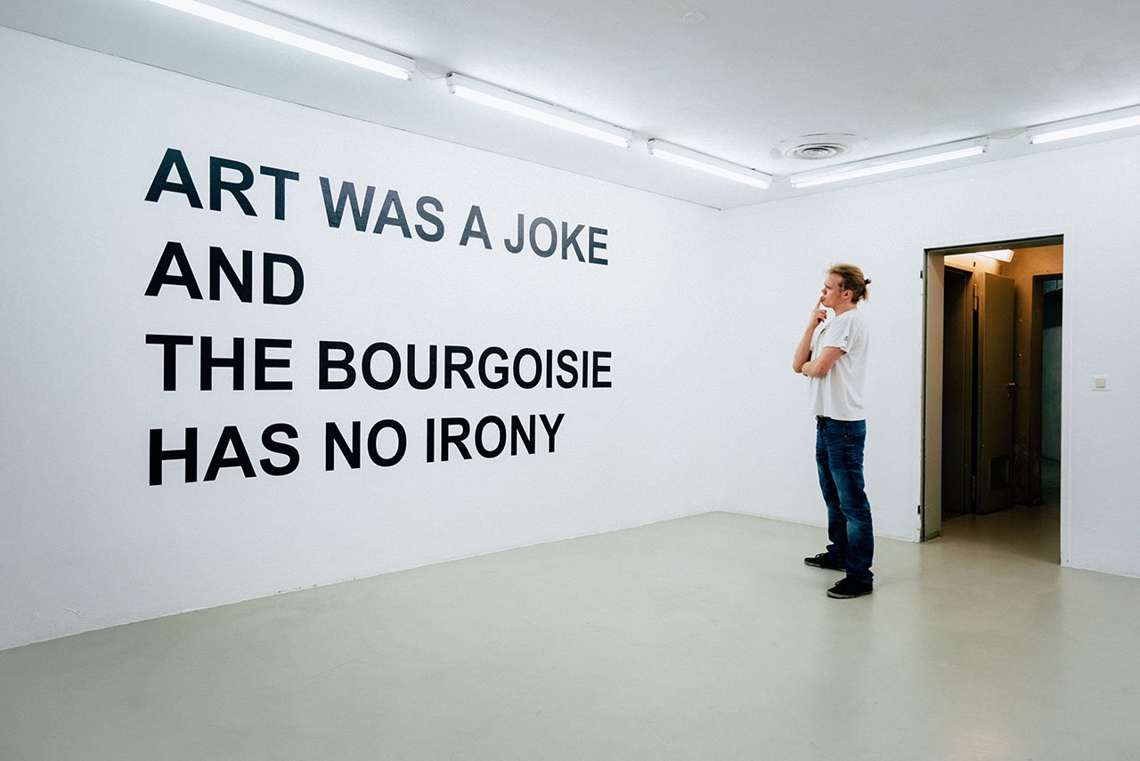 Wall text ART WAS A JOKE AND THE BOURGEOISIE HAS NO IRONY, a person standing in front