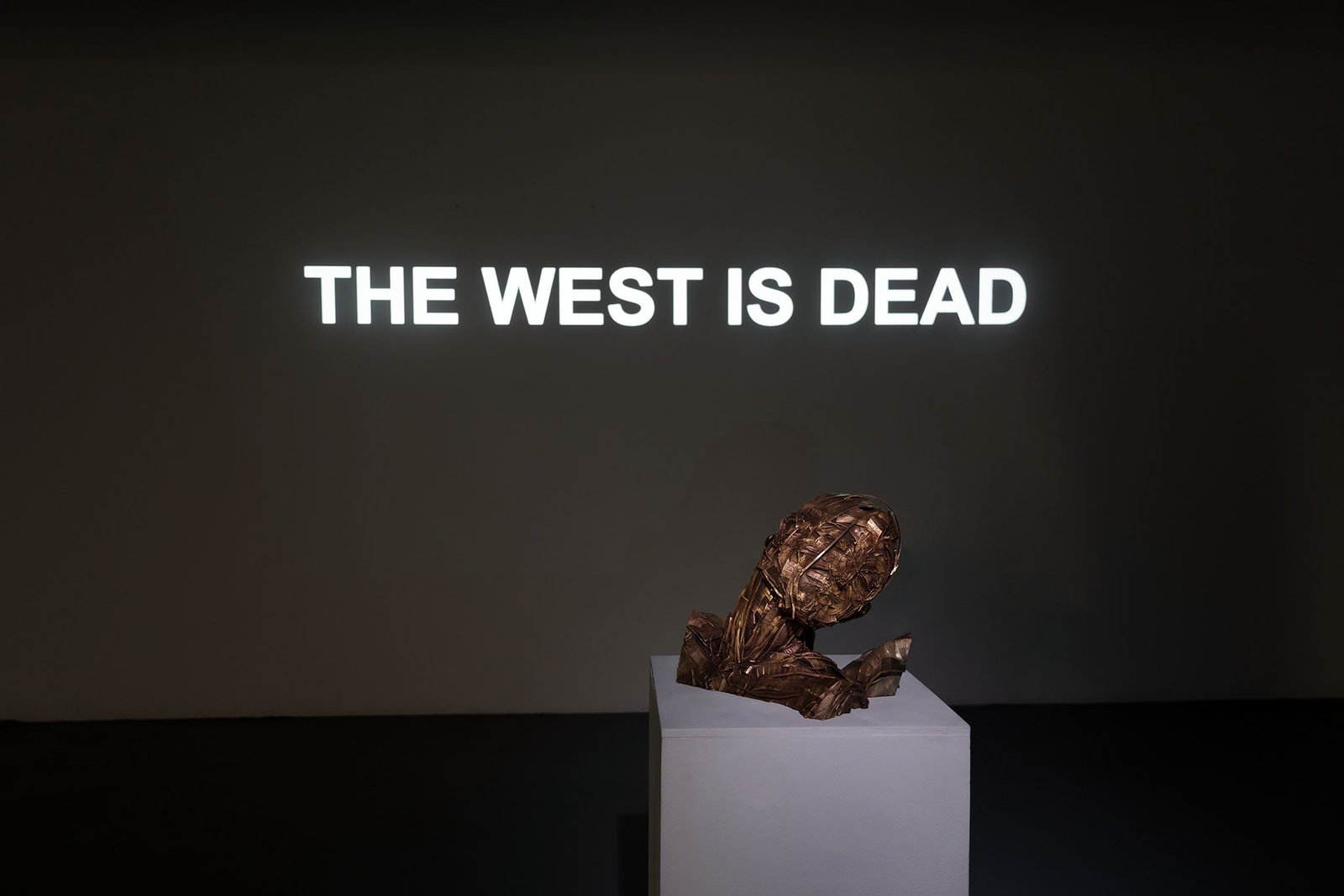 Bronze bust on a plinth, behind it THE WEST IS DEAD