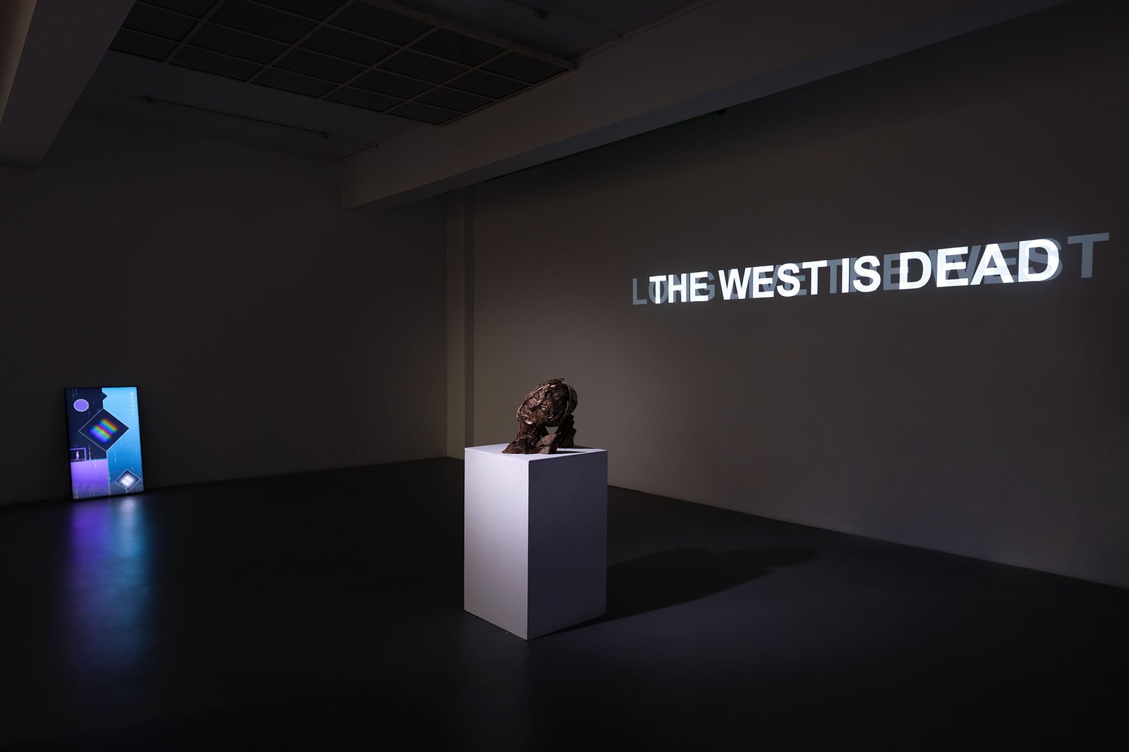 Darkened room with the projection THE WEST IS DEAD and the bronze bust on its plinth